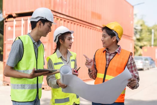 A diverse team of construction workers, including males and a female supervisor, reviews blueprints and shares ideas at a construction site surrounded by containers.