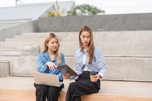 Two diverse women collaborating on a tablet and laptop while enjoying coffee in an outdoor workspace, emphasizing teamwork and productivity.