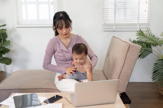 A caring mother sits at her workspace, focused on her laptop while playfully engaging with her baby, creating an inspiring balance of work and parenthood in a cozy home.