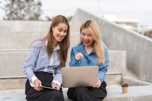 Two diverse women excitedly collaborating on a laptop and tablet, sharing ideas and insights in an outdoor setting.