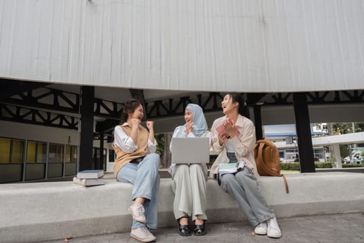 Diverse female students engaged in a lively conversation, smiling and sharing ideas while using a laptop and surrounded by books in a university campus setting.