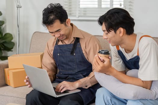 Two cheerful delivery professionals, one holding a coffee cup, happily reviewing orders on a laptop in a cozy office filled with packages.