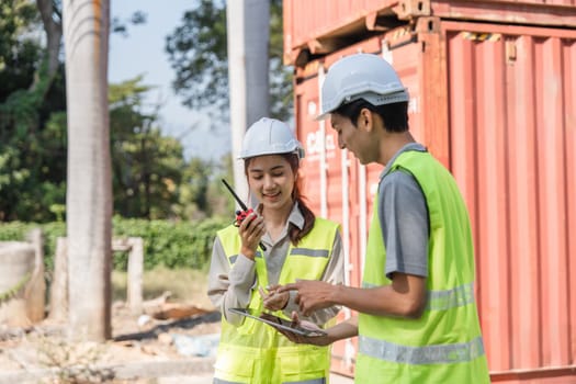 Two construction engineers use radios to communicate project updates while reviewing plans near shipping containers, emphasizing teamwork and coordination.