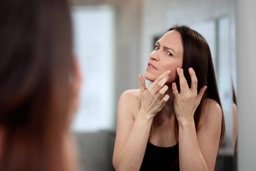 Anxious dermatologist examines acne on young woman's face in mirror.