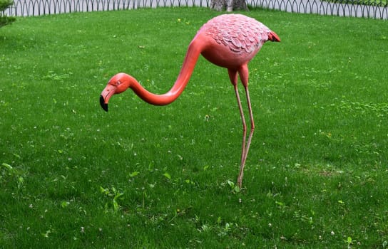 Kharkov, Ukraine, 14 May 2024. A figure of a pink flamingo on a green lawn. The figure has a long curved neck, a detailed pink body with feather texture, and slender legs. In the background, there is a fence and some bushes.