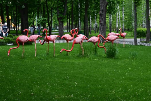Kharkov, Ukraine, 14 May 2024. A group of pink artificial flamingos in a park with green grass. The environment is surrounded by trees and people can be seen strolling in the background, indicating an outdoor leisure environment.