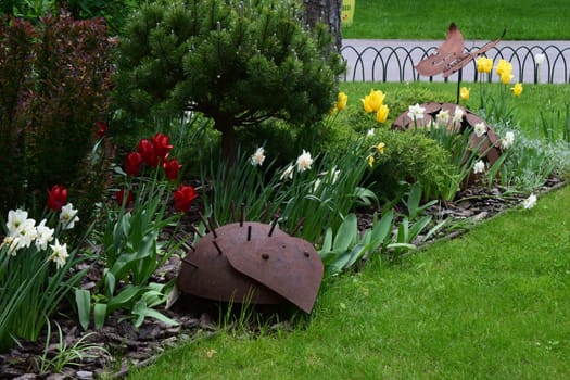 Kharkov, Ukraine, 14 May 2024. A garden decorated with metallic hedgehog sculptures. There are flowers of different colors, including red and yellow tulips and white daffodils. The surrounding grasses and shrubs are well tended, and a small paved path is visible in the background.