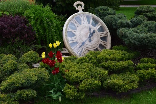 Kharkov, Ukraine, 14 May 2024. A large decorative pocket watch resting among bushes. The clock has Roman numerals and black hands. There are yellow and red flowers in the foreground along with lush green vegetation