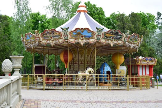 Kharkov, Ukraine, 14 May 2024. A vintage carousel with ornate decorations, featuring painted panels of scenic landscapes. The ride includes classic horses, a red car, and a blue rocket-shaped vehicle. Two large decorative spheres hang from the canopy, and the structure is enclosed by a yellow metal fence. The background shows green foliage, indicating an outdoor setting.