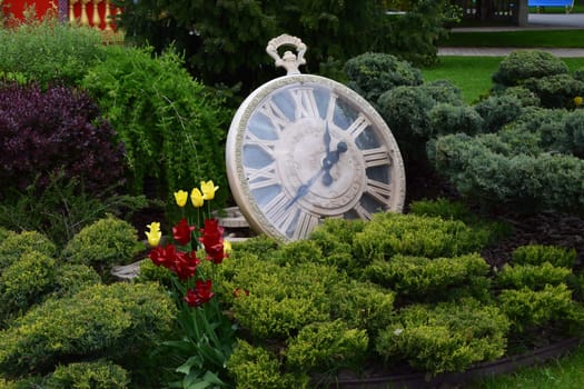 Kharkov, Ukraine, 14 May 2024. A large decorative pocket watch resting among bushes. The clock has Roman numerals and black hands. There are yellow and red flowers in the foreground along with lush green vegetation