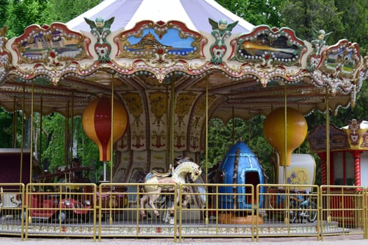 Kharkov, Ukraine, 14 May 2024. A vintage carousel with ornate decorations, featuring painted panels of scenic landscapes. The ride includes classic horses, a red car, and a blue rocket-shaped vehicle. Two large decorative spheres hang from the canopy, and the structure is enclosed by a yellow metal fence. The background shows green foliage, indicating an outdoor setting.