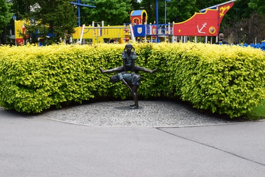 Kharkov, Ukraine, 14 May 2024. A statue of two children playing leapfrog, situated on a cobblestone circular base, surrounded by green shrubs. In the background, there is colorful playground equipment, a ship with vibrant yellow, blue, and red elements decorated with nautical symbols.