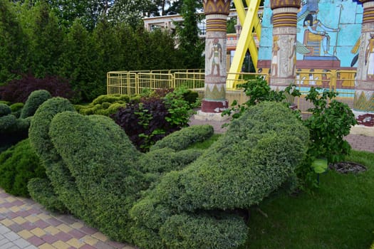 Kharkov, Ukraine, 14 May 2024. A garden scene with well-manicured greenery shaped into abstract topiary forms. In the background, there are ornate columns with Egyptian-style murals and decorations. The setting includes greenery, tiled pavement, and a colorful mural depicting Egyptian motifs.