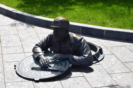 Kharkov, Ukraine, 14 May 2024. A bronze sculpture of a worker climbing out of a manhole. The worker is wearing a helmet and leaning on the manhole cover. The scene is set on a paved area with grass in the background. This artistic depiction may symbolize urban and underground labor.