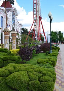 Kharkov, Ukraine, 14 May 2024. A park with a cobblestone path and well-kept gardens. On the left, a building with ornate architecture and light-toned details stands out. Next to the building, there are columns decorated with Egyptian motifs. In the center, there is a large red Ferris wheel with multicolored booths.