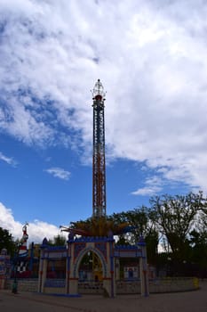 Kharkov, Ukraine, 14 May 2024. A carousel tower at an amusement park. The structure is vertical and multicolored, with rich decoration at the base. The sky is partially obscured by clouds, with blue spots visible. The environment is green, with trees visible in the background.