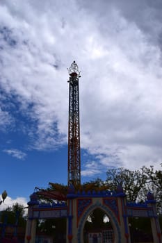 Kharkov, Ukraine, 14 May 2024. A carousel tower at an amusement park. The structure is vertical and multicolored, with rich decoration at the base. The sky is partially obscured by clouds, with blue spots visible. The environment is green, with trees visible in the background.
