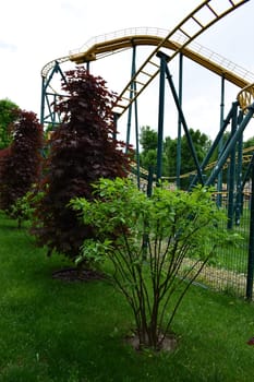 Kharkov, Ukraine, 14 May 2024. A roller coaster of metal structure with yellow rails and dark green support. It is set in an environment with green grass and trees around it. The sky is overcast, indicative of a partly sunny day.