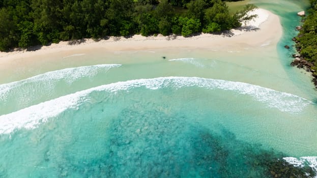 Waves crash against rocks at a lush green coastline, surrounded by turquoise waters. Seychelles, Mahe. Grand Anse.