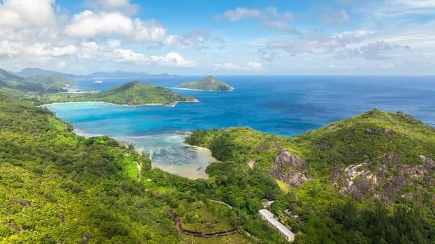 Coastal landscape with turquoise bays, lush green hills and distant islands under a cloudy sky. Seychelles, Mahe.