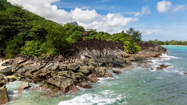 Rocky coastline with lush greenery and turquoise waters under a partly cloudy sky. Seychelles.