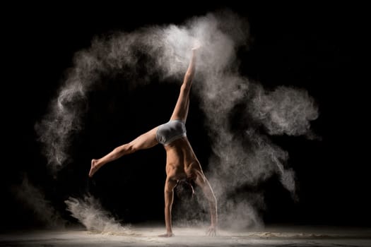 Back view of anonymous male dancer performing handstand with split in circle of dust against black background