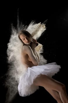 Side view of youthful soft topless woman in cloud of white powder with closed eyes wearing white ballet tutu on black background in studio