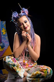 Sexy girl with bright make up and pink braids sitting on the floor near easel view against black background