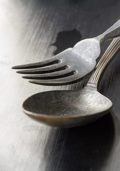 The morning sun shines hits on Ancient worn vintage cutlery on the black background. Close up of Crossed black metal spoon and fork, Highlighting its texture and design, Light and shadow, use it as your Space for text, Design, Wallpaper and Poster, Selective focus.