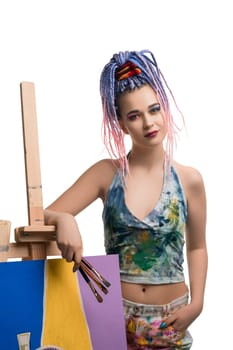 Stylish young female in colorful dirty clothes looking at camera and leaning on easel with painting against white background