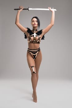 Full body sexy woman in armor shaped underwear stepping forward and carrying sharp sword in raised arms against gray background