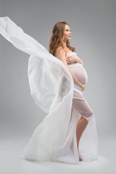 Full body young lady wrapped in thin white cloth touching pregnant belly against gray background