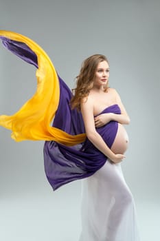 Beautiful pregnant female covering breast with waving colorful fabric and touching tummy against gray background