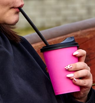 A young woman with long brown hair and white headphones is drinking from a pink cup with a straw. She is wearing a black sweatshirt and has her nails painted pink with white polka dots.