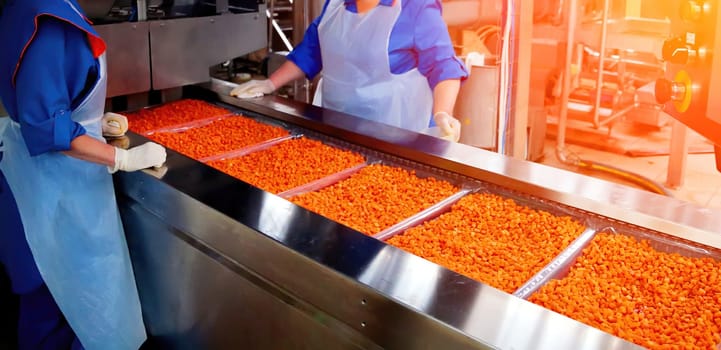 Two employees in blue uniforms and aprons work diligently at a snack production line, handling vibrant orange snacks ready for packaging in a factory.