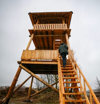 A person climbs the wooden steps of a tall observation tower set in a natural landscape, under overcast skies, ready to enjoy the view.