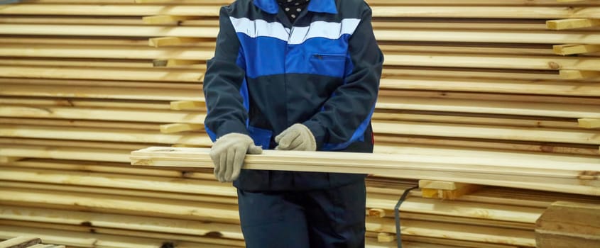 A worker in blue and black attire uses a measuring tape on a wooden plank in a busy lumber yard filled with stacked timber.
