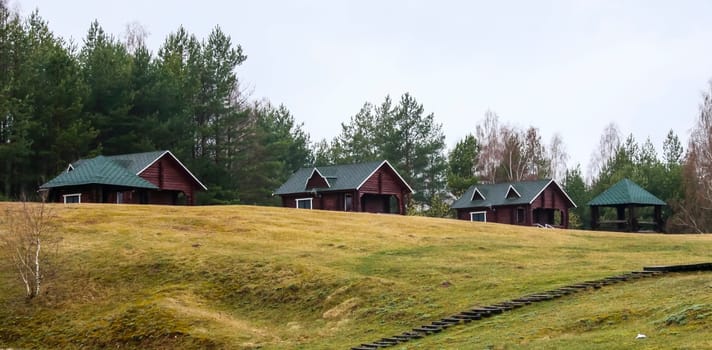 Several wooden cabins are situated on a grassy hillside, surrounded by trees under a cloudy sky, creating a peaceful retreat atmosphere.