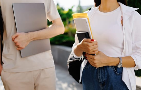 Anonymous young male with laptop and unrecognizable female with books in casual clothes while standing together on street in sunlight against blurred green trees park and blue sky