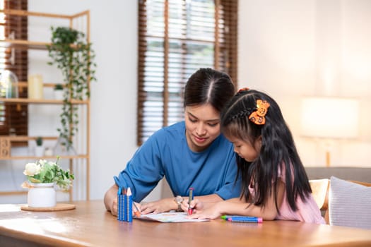 A young family spends their free time together in the living room at home. Mother and little daughter draw pictures with crayons on paper, smiling happy..