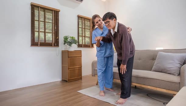 A caring young Asian nurse helps an elderly person walk in the house. An elderly disabled man tries to walk with the help of a female caregiver..