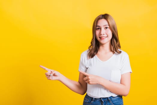 Asian Thai happy portrait beautiful cute young woman standing wear white t-shirt pointing finger away side looking to camera, studio shot isolated on yellow background with copy space