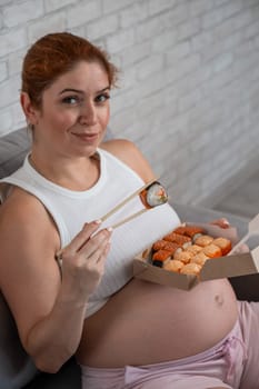 A pregnant woman sits on the sofa and eats rolls from a box. Food delivery