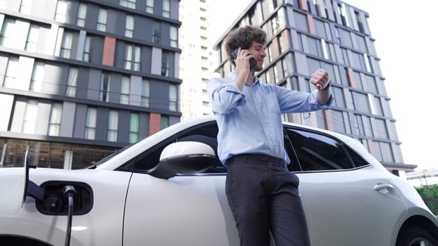 Suit-clad businessman with progressive ambition leaning on his electric vehicle while standing on a charging station with a power cable plug and a renewable energy-powered electric vehicle.