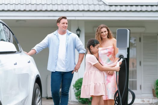 Happy little young girl learn about eco-friendly and energy sustainability as she help her family recharge electric vehicle from home EV charging station. EV car and modern family concept. Synchronos