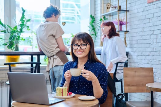 Middle aged woman in bakery cafeteria with cup of coffee and dessert cake, sitting at table, with laptop, happy smiling female looking at camera. Lunch break, food, lifestyle, mature 40s people concept