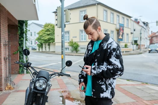 Young trendy male with phone and an electronic cigarette standing on motorcycle on city street. Youth, lifestyle concept