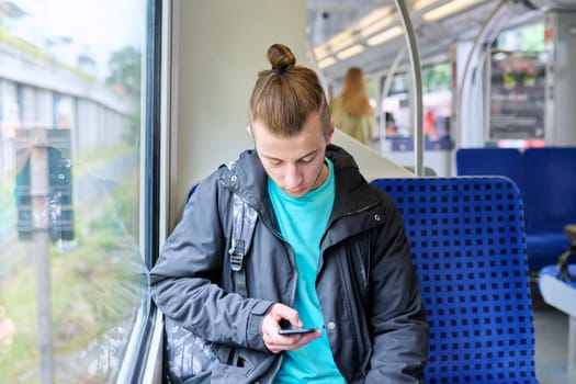Young male passenger sitting inside an electric city commuter train. Guy student with backpack using smartphone, lifestyle, youth, railway transport concept.