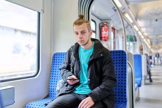 Young male passenger sitting inside an electric city commuter train. Guy student with backpack using smartphone, lifestyle, youth, railway transport concept.