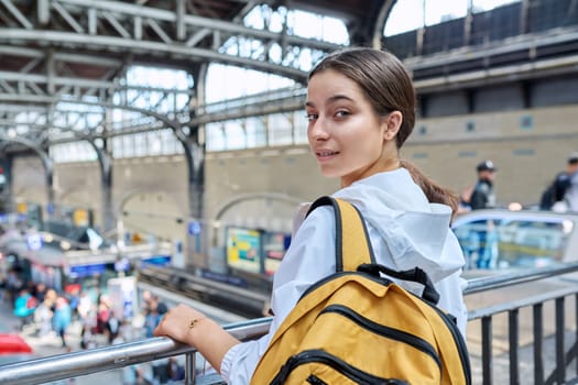 Teenage girl with backpack looking at camera at railway station. High school student, teenager girl passenger, suburban urban and intercity rail transport